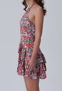 Kukallinen halterneck-mekko, jossa on röyhelöhelma. Valmistettu kevyestä kankaasta, jossa on sininen tausta ja eloisat vaaleanpunaiset, punaiset ja vihreät kukkakuviot.