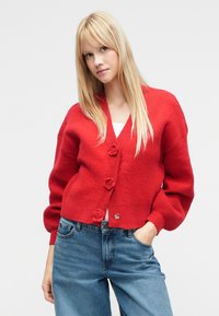 Sarkans adīts kardigans ar V veida kakla izgriezumu, trim dekoratīvām rožu pogām un piepūstām piedurknēm, saskaņots ar gaiši ziliem džinsa džinsiem.
