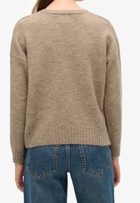 Beżowy sweter z ribbowym wykończeniem i okrągłym dekoltem, o luźnym kroju i fakturowanym splocie. W połączeniu z niebieskimi dżinsami.