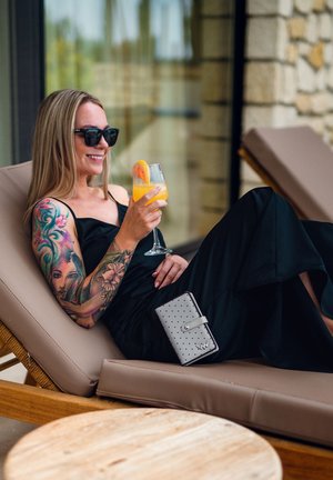 Black sleeveless dress, clear glass of orange drink with garnish, gray wallet with perforated pattern, relaxed on a tan lounge chair.