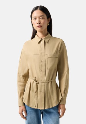 Frau trägt eine beigefarbene, langärmelige Hemdbluse mit Taillenband und blaue Jeans, steht vor einem schlichten Hintergrund.