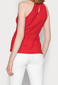 Top rojo sin mangas con una espalda con abertura, tela texturizada y detalle de cintura fruncida, combinado con pantalones blancos ajustados.