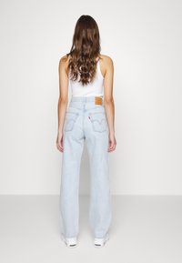 Jeans de pierna ancha de color azul claro con una cintura alta, cuentan con bolsillos traseros y un parche de cuero marrón en la cinturilla. Llevados con una camiseta sin mangas blanca.