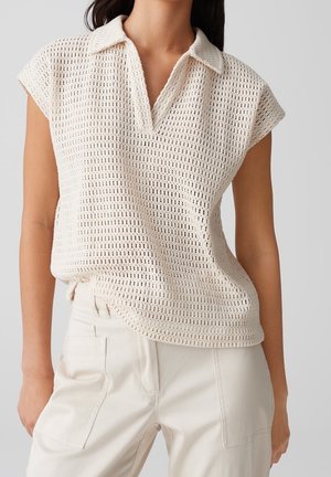 Femme portant un haut en tricot crocheté crème sans manches avec un col en V et un pantalon taille haute écru.