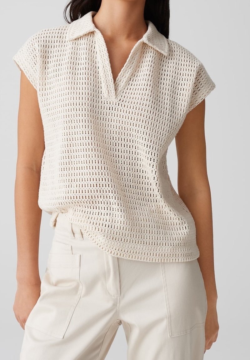 Femme portant un haut en tricot crocheté crème sans manches avec un col en V et un pantalon taille haute écru.