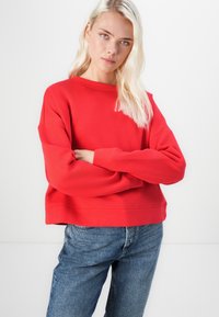 Sudadera recortada roja con un escote redondo y costuras de hombro caídas, combinada con vaqueros de denim azul. Textura suave, diseño casual.
