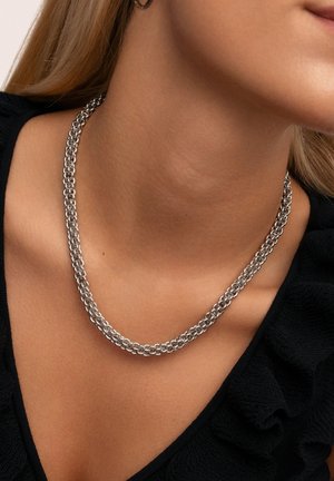 Collier en chaîne argentée avec des maillons entrelacés, profil plat et texture lisse, porté autour du cou, associé à un haut noir à volants.