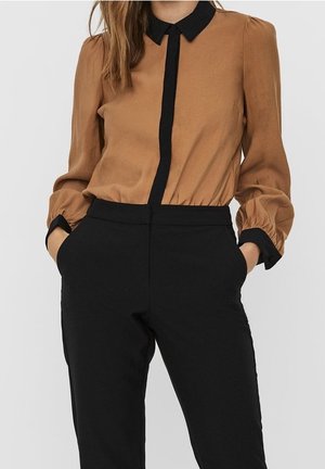 Vrouw met een bruin blouse met lange mouwen, zwarte kraag en manchetten, gecombineerd met hooggetailleerde zwarte broek, handen in de zakken.