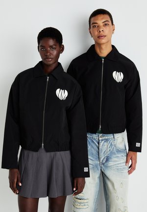 Svarte cropped jakker med glidelås foran, med hvite hjerte-logger på brystet. En jakke har elastisk kant, og er kombinert med et grått plisserte skjørt og slitte jeans.
