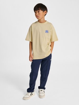 Junge steht mit den Händen in den Taschen, trägt ein beiges oversized T-Shirt, marineblaue Jogginghosen und weiße Sneakers mit schwarzen Streifen.