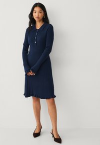 Dunkelblaues Rippstrickkleid mit langen Ärmeln, Knopfleiste und ausgestellt gerafftem Saum. Glatte Textur, figurbetonte Silhouette und Kragen-Details.