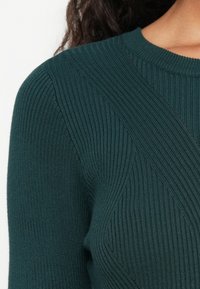 Gros plan sur un pull en maille côtelée bleu sarcelle foncé avec des motifs texturés diagonaux et verticaux sur l'épaule et la poitrine.