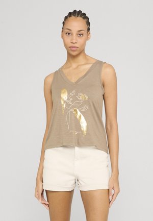 Kvinde med flettet hår iført beige ærmeløs top med gyldent abstrakt design og lyse shorts mod en ensfarvet baggrund.