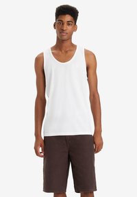 Top in cotone bianco senza maniche con scollatura rotonda, abbinato a pantaloni corti marroni. Il top presenta un piccolo logo vicino al petto.