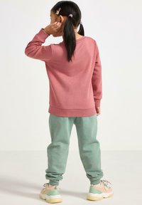 Barn med mørkt hår i hestehaler iført en lyserød sweatshirt, grønne sweatpants og multifarvede sneakers, stående med ryggen til, hånd tæt på øret.
