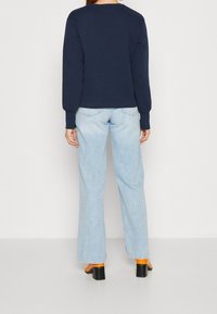 Felpa a maniche lunghe di colore blu navy con scollo rotondo, abbinata a jeans a gamba larga di un blu chiaro. Scarpe con tacco nere con accenti arancioni.