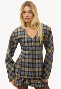 Femme blonde portant un blazer structuré à carreaux marron, noir et bleu avec des épaules bouffantes et une mini-jupe assortie à volants.