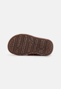 Semelle de chaussure en caoutchouc brun avec des motifs de grip texturés et des détails hexagonaux. Forme légèrement incurvée avec des bords surélevés et logo de la marque.