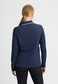 Polo a maniche lunghe blu navy con colletto bianco. Presenta una texture liscia e un logo sulla manica sinistra. La vista posteriore mette in evidenza una vestibilità sartoriale.