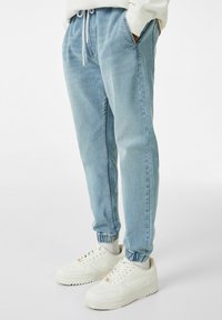 Light blue denim joggers with an elastic waistband, side pockets, and elastic cuffs, paired with white sneakers. Smooth texture, casual design.