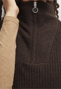 Haut sans manches en maille côtelée marron avec un col montant, doté d'une fermeture éclair argentée. Superposé à une chemise à manches longues marron clair.