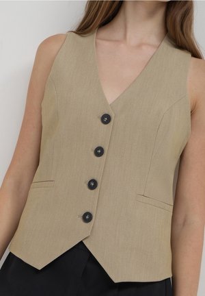 Veste beige sans manches avec quatre boutons noirs portée sur un pantalon noir, montrant le haut du torse et le cou d'une personne aux longs cheveux.