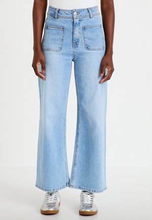 Personne portant un jean large bleu clair avec de grandes poches avant et des baskets argentées, debout devant un fond uni.