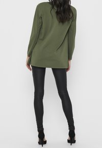 Top de manga larga verde oliva con un ajuste relajado y dobladillo redondeado, combinado con leggings ajustados negros, complementado con zapatos de tacón alto negros.