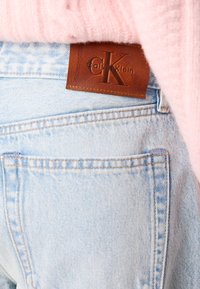 Light blue denim jeans with visible stitching and pockets, featuring a dark brown leather tag embossed with "Calvin Klein." Soft pink fabric above.