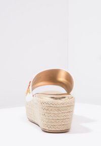 Sandale à plateforme blanche avec une base en jute tressé. Accents dorés sur le talon et l'arrière. Présente un design incurvé et des ornements floraux.