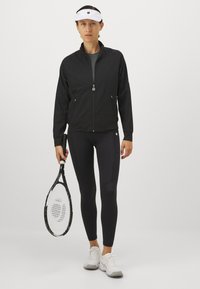 Naine mustas sportlikus jakis ja retuusides, hoides tennisereketit, kandes valget visorit ja valgeid jooksujalatseid, seismas ühevärvilise tausta ees.