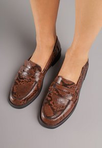 Mocassins en cuir avec imprimé serpent marron, présentant une texture lisse, une large sangle sur le devant et une conception de bout arrondi.