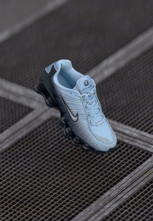 Baskets Nike bleu clair avec semelle noire et logo, flottant au-dessus d'une surface en grille métallique, vues d'un angle supérieur incliné.