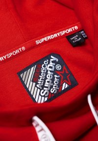 Interior de sudadera roja con capucha que presenta una etiqueta negra tejida con texto blanco y rojo. La etiqueta incluye un logotipo y la información de cuidado. Textura suave.