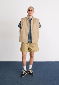 Colete bege sem mangas, camisa às riscas, calções caqui, meias brancas, ténis pretos; usado por uma pessoa com cabeça rapada e óculos de sol.