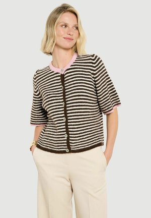 Blonde Frau trägt eine braun-weiß gestreifte Kurzarm-Strickjacke mit pinkem Besatz, beige Hose und ein goldenes Armband am linken Handgelenk.