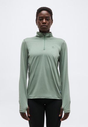 Fekete személy világoszöld, hosszú ujjú cipzáros felsőt és fekete nadrágot visel, szemben áll egy egyszerű fehér háttér előtt.