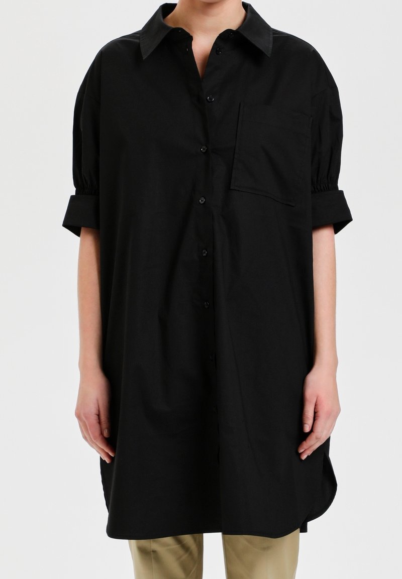 Chemise noire ample à boutons avec manches courtes retroussées et poche poitrine portée sur un pantalon beige, présentée du cou jusqu'à la mi-cuisse.