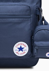 Sac à dos en tissu navy avec plusieurs poches, présentant un logo Converse blanc rond avec une étoile bleue. Textures robustes et fermetures éclair visibles.