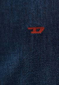 Κοντινή λήψη σκουρότερου μπλε υφάσματος τζιν με μικρά κόκκινα κεντημένα γράμματα "JD" στην επιφάνεια.