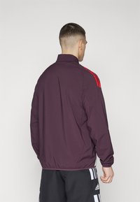Bordeaux sportsjakke med høy krage, elastiske mansjetter og hem. Har tre røde striper på skulderen og en glatt tekstur.