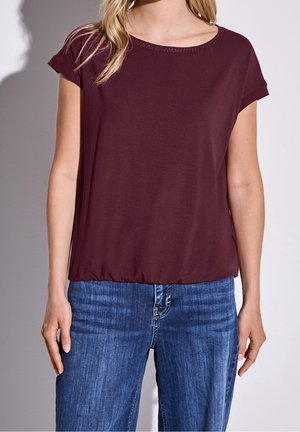 Bordeauxrood T-shirt met korte mouwen en een ronde hals, met subtiele details langs de kraag, gecombineerd met een blauwe spijkerbroek.