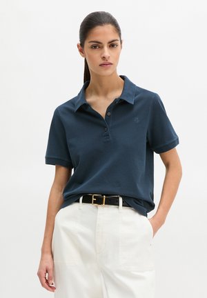 Junge Frau mit dunklem Haar, trägt ein marineblaues Polohemd, das in weiße Hosen mit einem schwarzen Gürtel gesteckt ist, steht mit einer Hand in der Tasche.