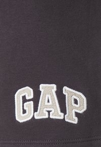 Tmavě šedá tkanina s hladkým povrchem, se velkými bílými vyšívanými písmeny "GAP" v sportovním písmu.