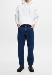 Sudadera blanca con capucha y bolsillo delantero, combinada con jeans de corte suelto en azul oscuro y botas negras de tobillo. Diseño sencillo y colores sólidos.