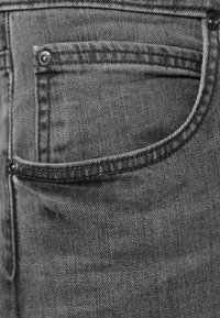 Jeans in denim grigio presentano una tasca frontale con un bottone in metallo e cuciture a vista, mostrando una texture morbida e una leggera sbiaditura.