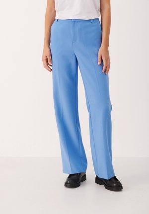 Personne portant un pantalon large bleu clair, un haut blanc et des mocassins noirs épais, debout devant un fond clair uni.