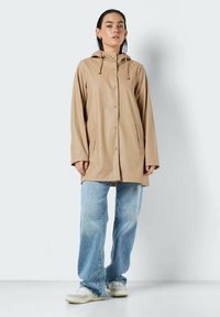 Manteau imperméable beige avec une capuche, des boutons-pression et des poches latérales. Associé à un jean bleu clair à jambes larges et des baskets blanches.