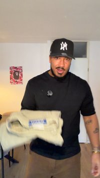 Μπεζ παντελόνι κρατημένο στο χέρι, κατασκευασμένο από ύφασμα με διπλωμένο σχέδιο και εμφανές ετικέτα. Μαύρο μπλουζάκι και καπέλο με λογότυπο. Απλό εσωτερικό φόντο.