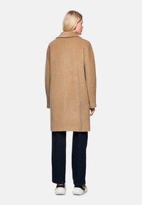 Beige Wollmantel mit breitem Kragen, langen Ärmeln und geradem Schnitt, getragen über dunklen Jeans und weißen Sneakers. Rückansicht sichtbar.